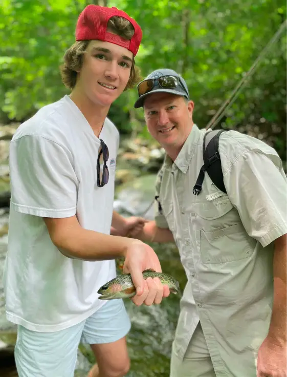 A young man in a red hat posing for a photo while holding a small fish and standing next to a man wearing a backpack.